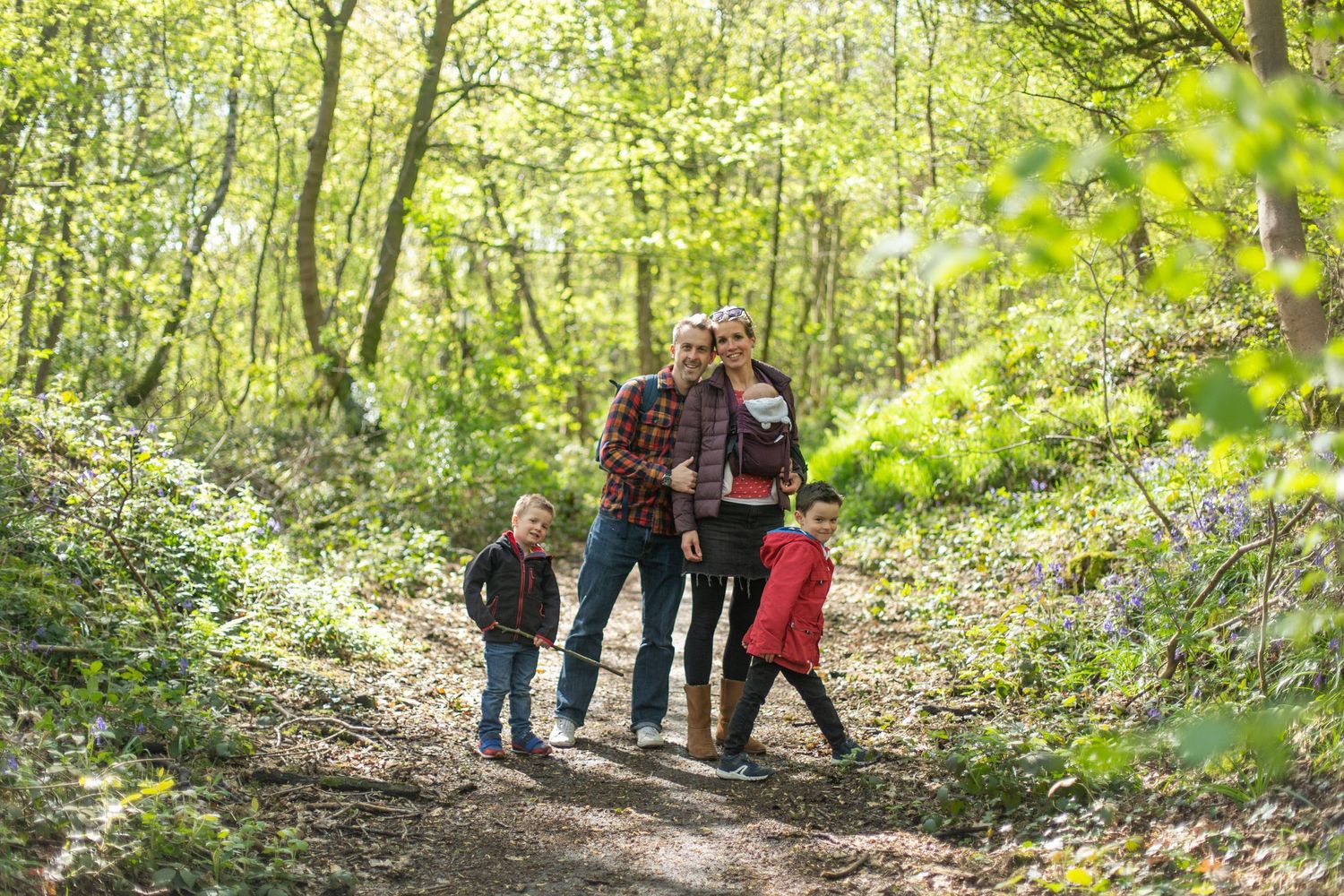 A group wearing plaid and casual attire walks together on a sunlit forest trail surrounded by lush green foliage.