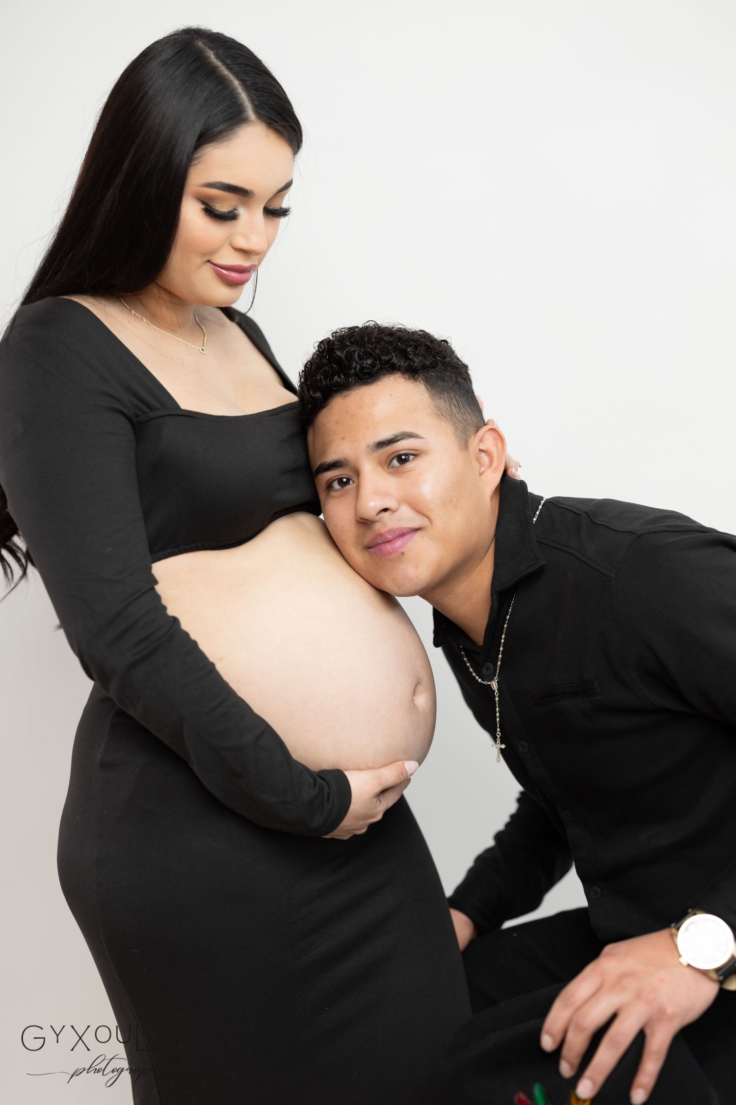 Maternity photo of couple in black outfits posing together against white background.