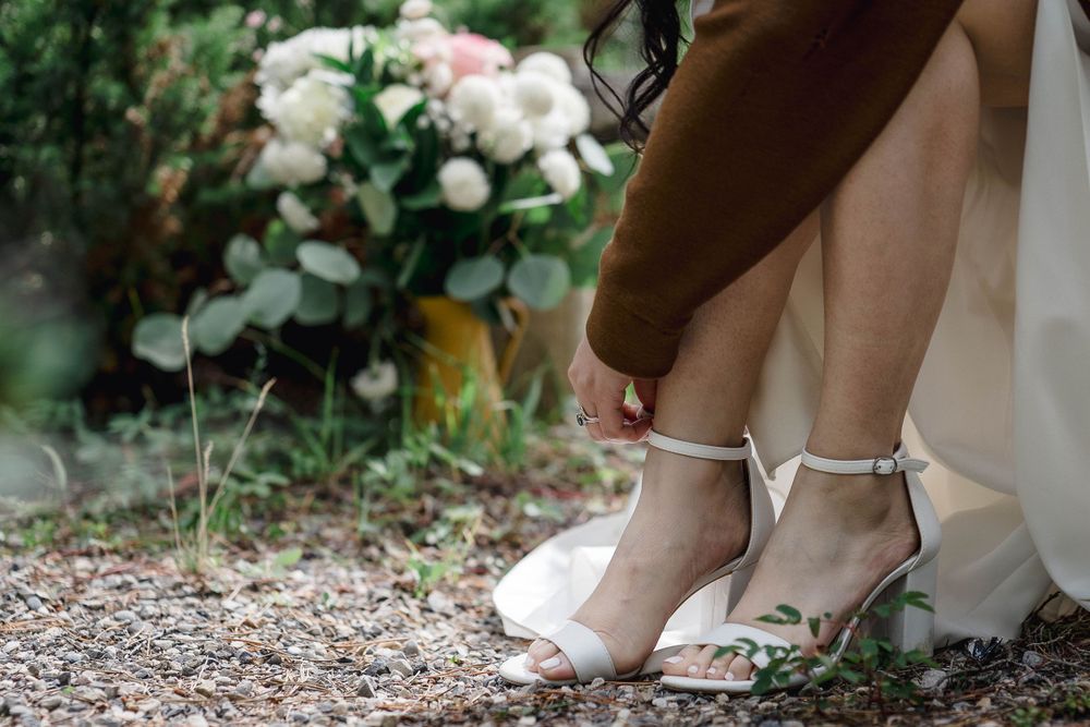 Elegant white bridal shoes with delicate ankle straps shown on a gravel path with white roses in the background.