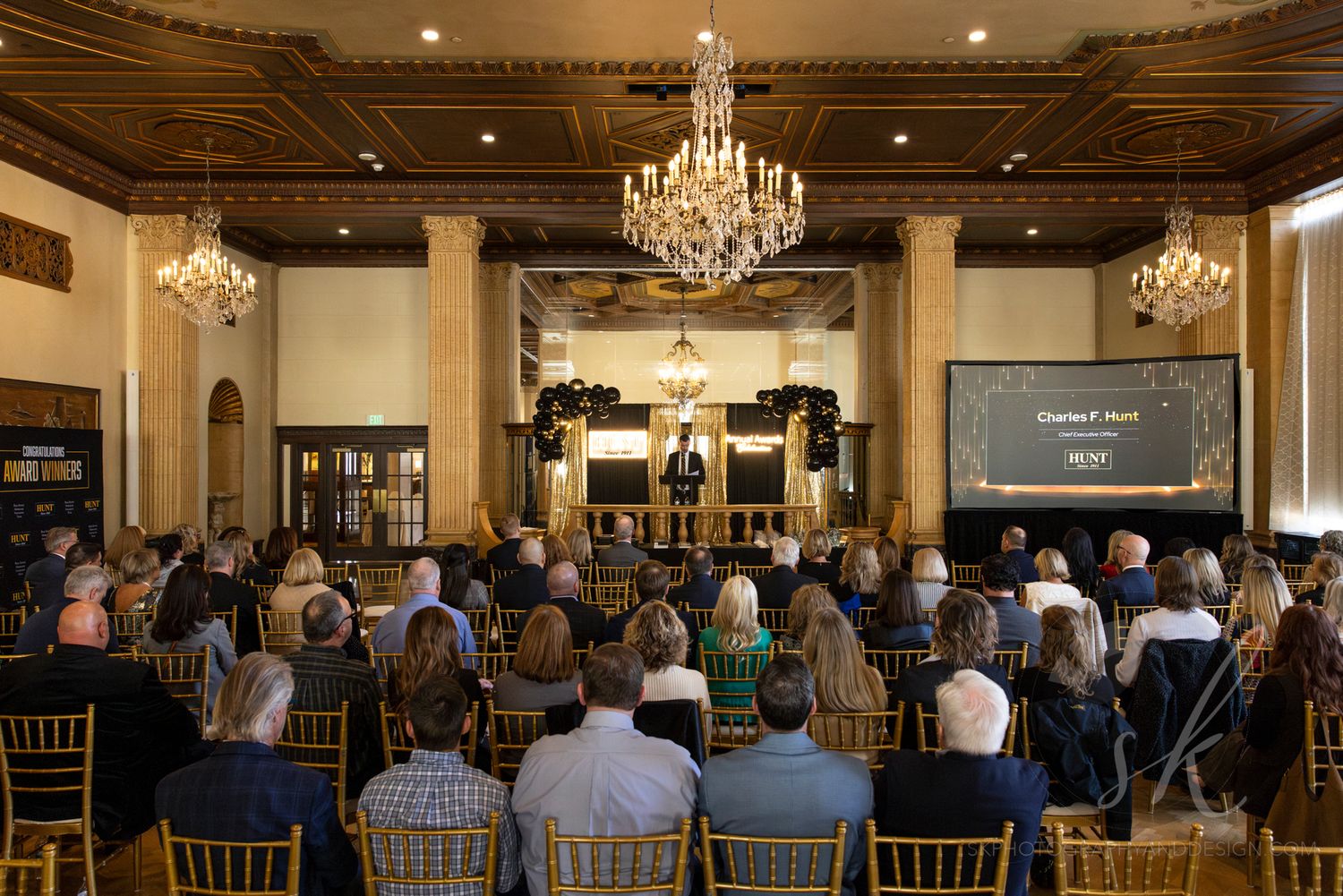 Elegant ballroom event with audience seated in gold chairs facing a presenter at a podium under crystal chandeliers.