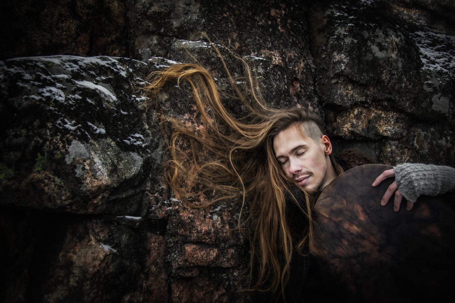 Dark moody portrait against stone wall shows figure with long flowing hair in dramatic lighting and shadowy atmosphere.