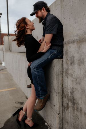 A couple shares an intimate moment against a concrete wall, with her in black dress and him in denim and baseball cap.