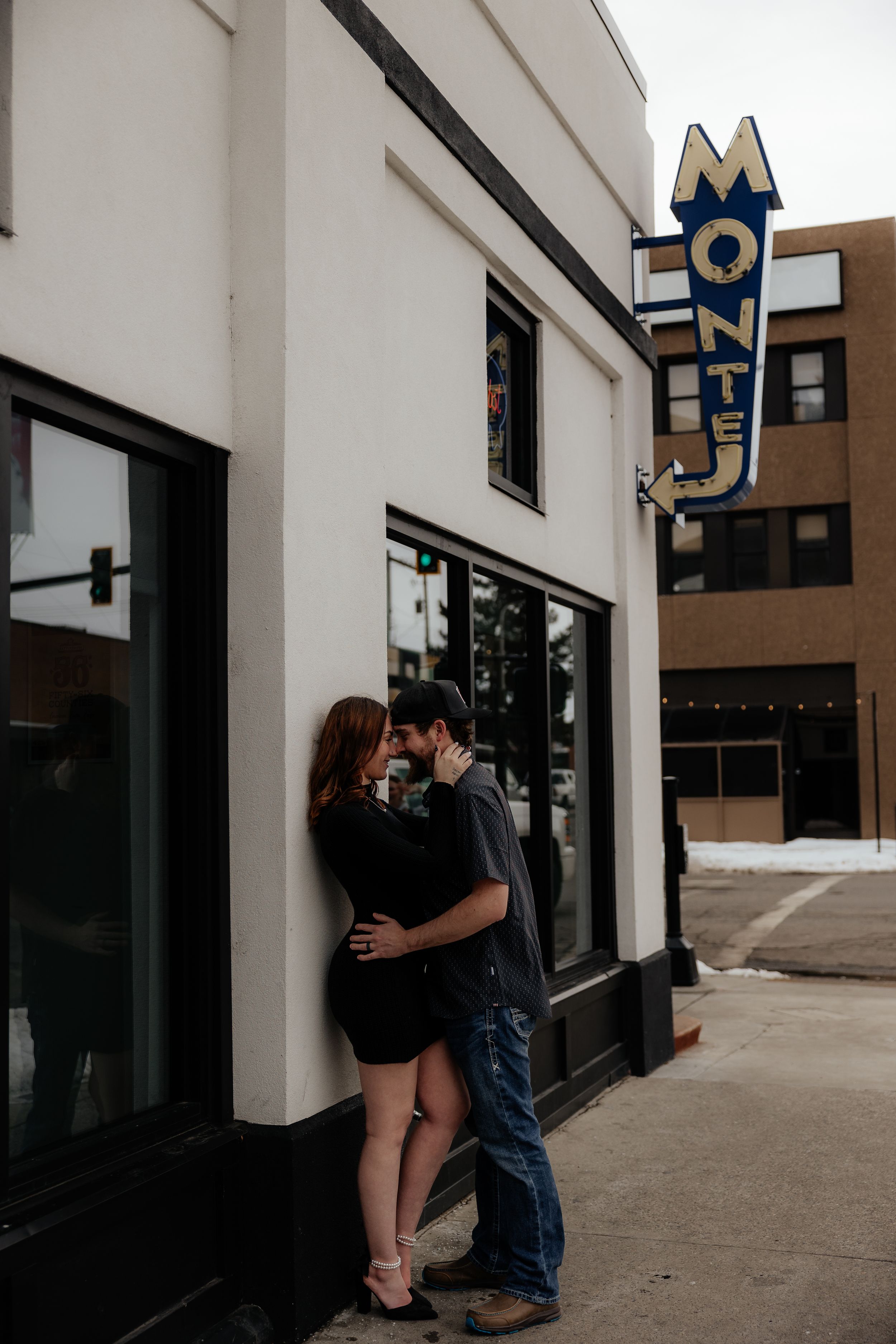 A couple embraces against a white building wall near a vintage motel sign in an urban setting.