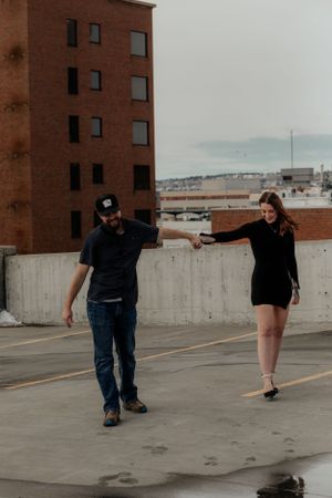 Two people walking casually on a rooftop parking deck with brick buildings and a cloudy skyline in the background.