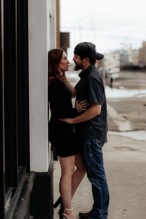 A romantic couple embraces against a wall outdoors, with her in a black dress and him in jeans and a baseball cap.