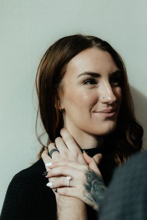 A woman smiling warmly while showing her engagement ring against a light wall.