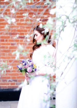 Elegant bride in white gown holds a colorful bouquet beside a brick wall framed by spring blossoms.