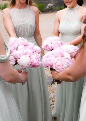 Two bridesmaids in sage green halter gowns hold lush pink peony bouquets at an outdoor wedding.