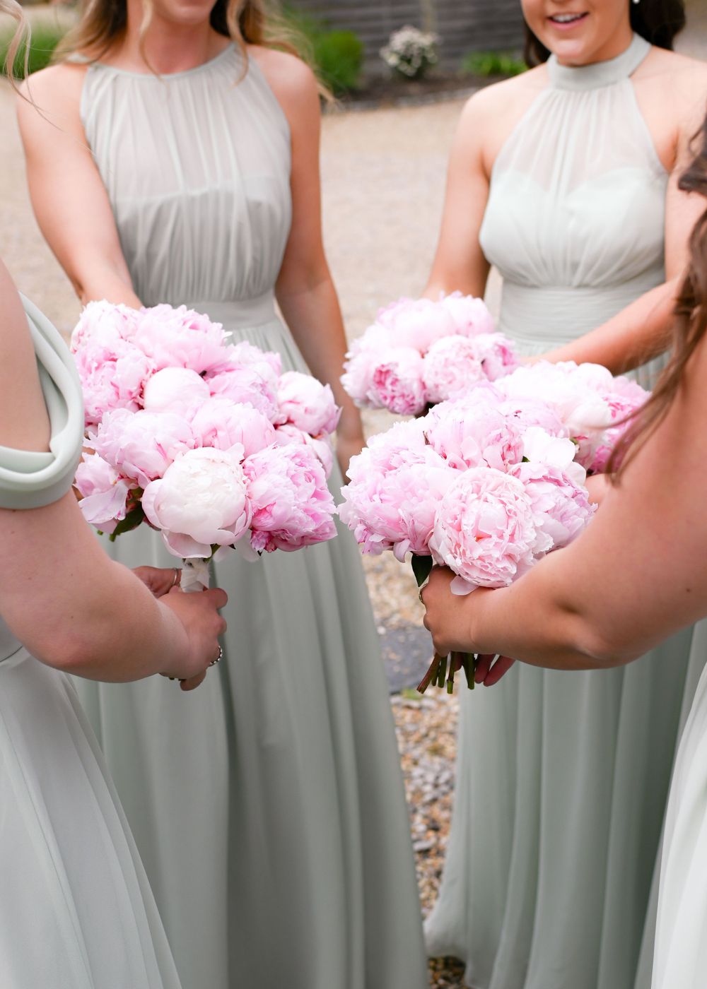Two bridesmaids in sage green halter gowns hold lush pink peony bouquets at an outdoor wedding.
