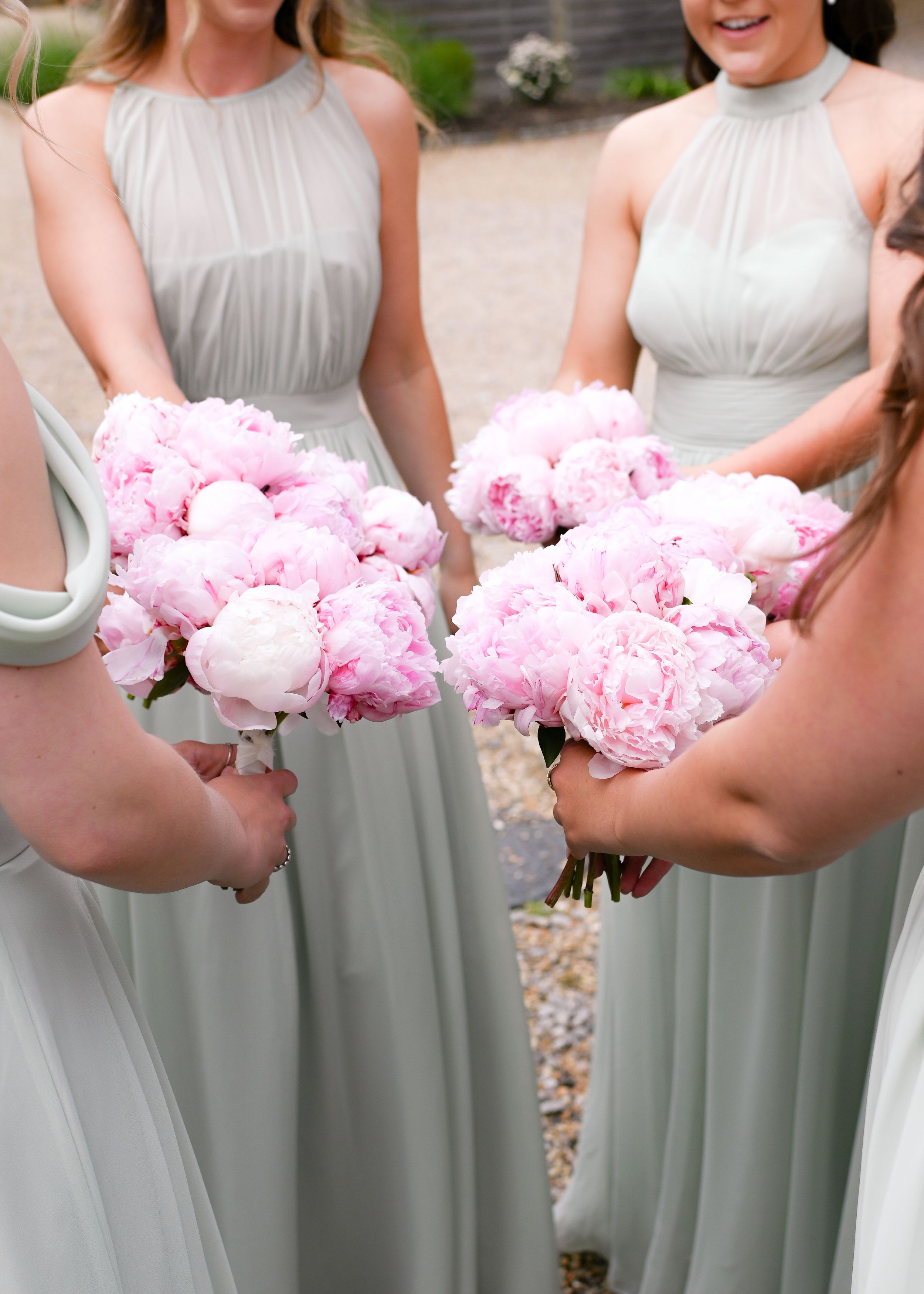 Two bridesmaids in sage green halter gowns hold lush pink peony bouquets at an outdoor wedding.