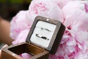 Two wedding rings displayed in a wooden ring box surrounded by soft pink peony flowers.