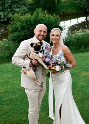 Smiling newlyweds pose outdoors holding a pug in a bow tie and a colorful wedding bouquet.