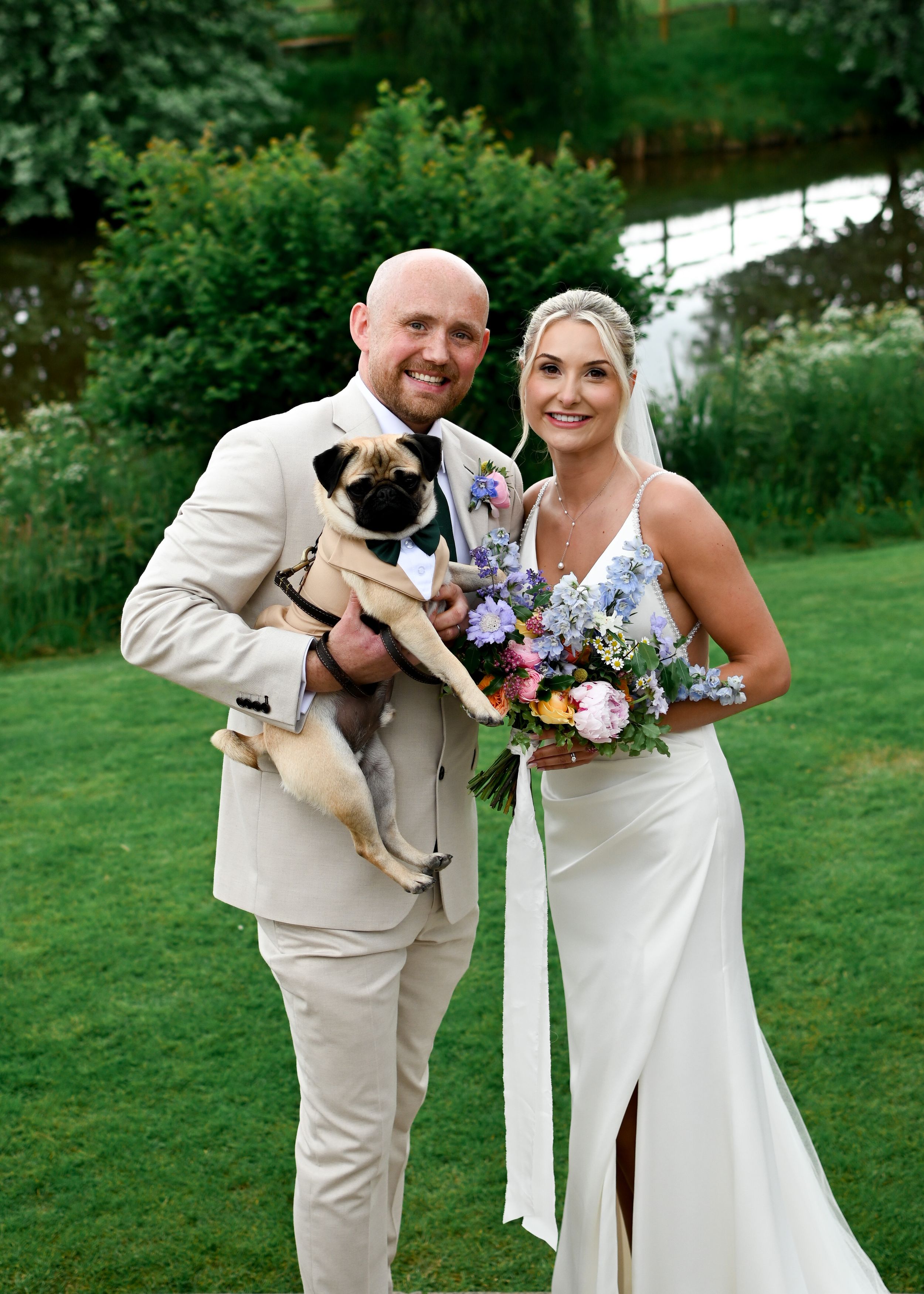 Smiling newlyweds pose outdoors holding a pug in a bow tie and a colorful wedding bouquet.