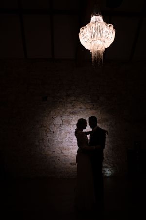 Romantic silhouette of wedding couple embracing beneath a glowing crystal chandelier against a stone wall backdrop.