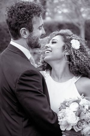 Black and white photo of interracial wedding couple gazing at each other, bride holding bouquet with flower in curly hair.