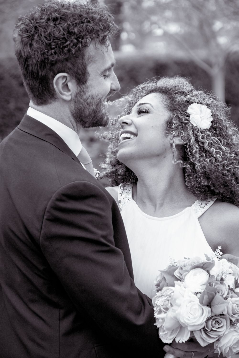 Black and white photo of interracial wedding couple gazing at each other, bride holding bouquet with flower in curly hair.