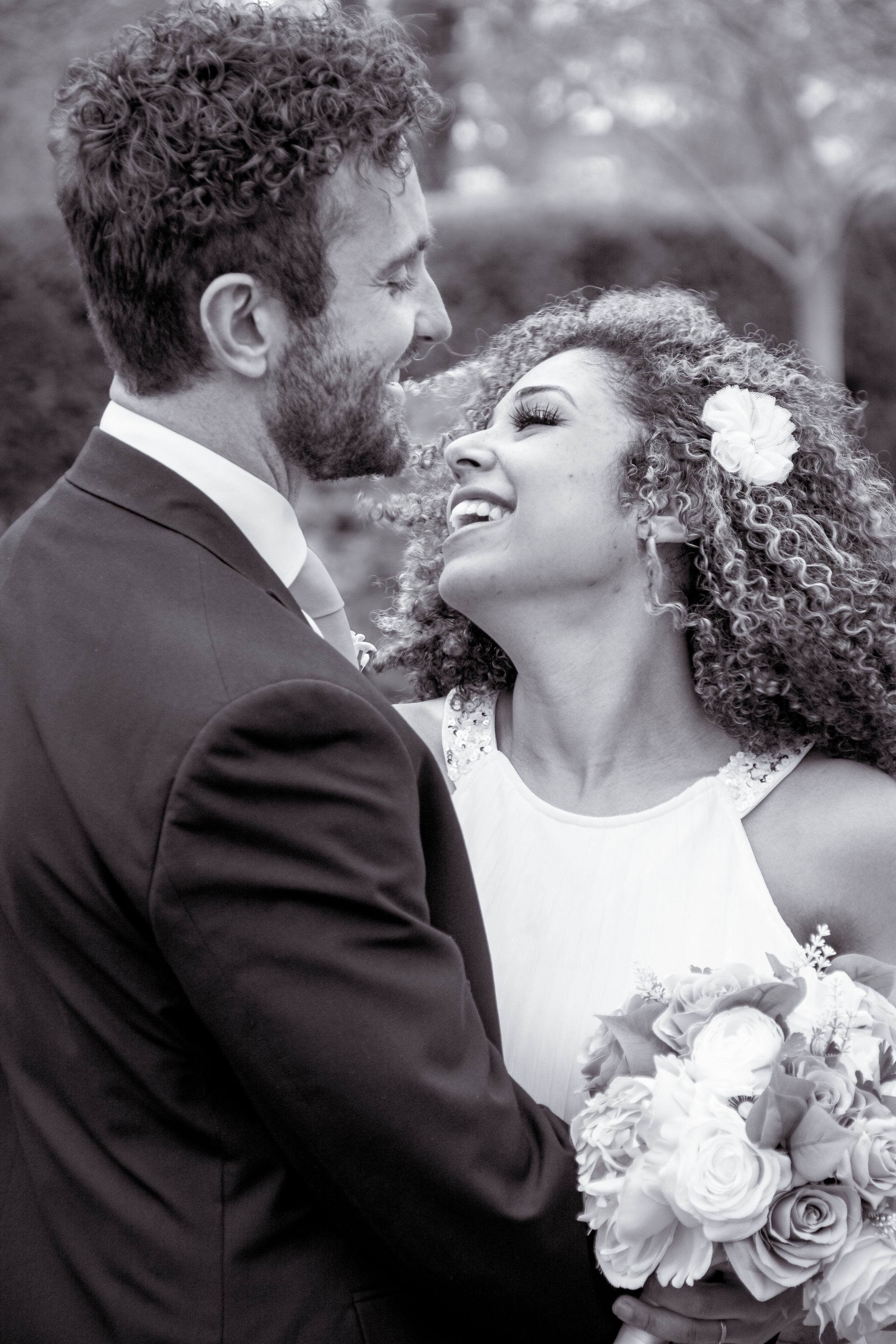 Black and white photo of interracial wedding couple gazing at each other, bride holding bouquet with flower in curly hair.