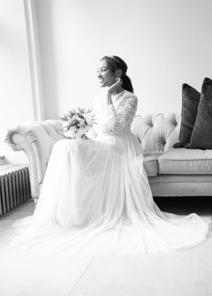 Black and white portrait of bride in lace long-sleeve gown seated on tufted sofa holding bouquet by bright window.
