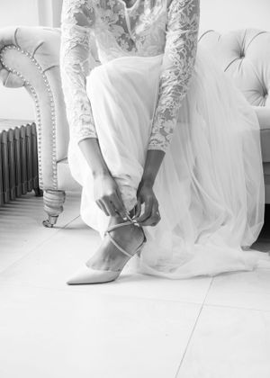 Black and white close-up of bride in lace wedding dress tying ribbon on pointed heels while seated on tufted chair.