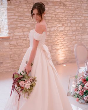 Elegant bride in off-shoulder ball gown holding colorful bouquet standing before a rustic stone wall at wedding venue.