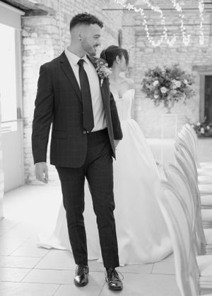 Black and white photo of stylish groom in dark plaid suit walking with bride in strapless gown at wedding venue.