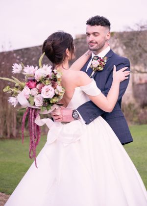 Romantic outdoor wedding portrait of couple embracing, bride in off-shoulder gown holding pink bouquet, groom in navy suit.