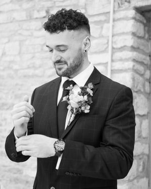 Black and white portrait of dapper groom in plaid suit adjusting cufflinks with floral boutonniere and luxury watch.