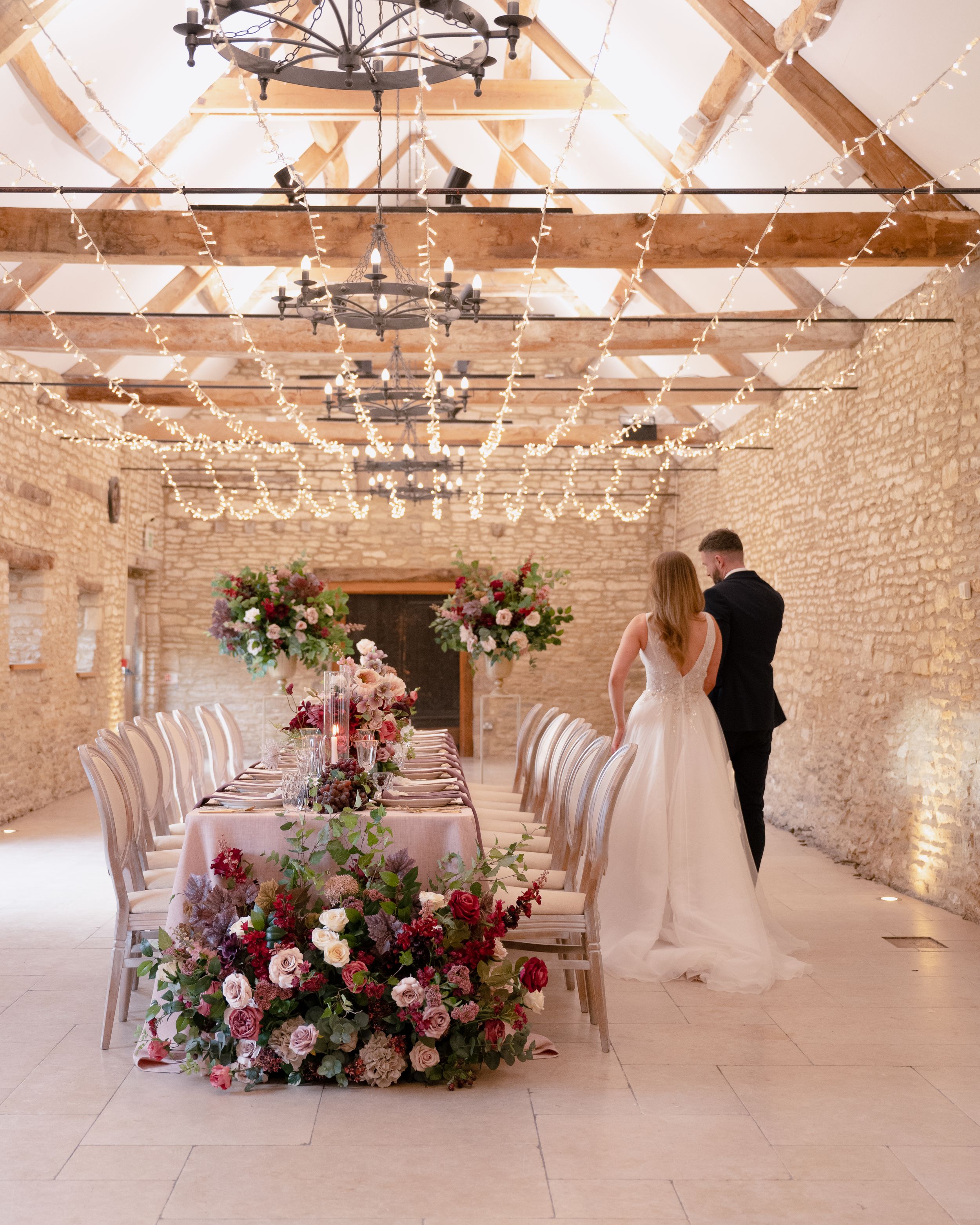 Bride and groom admire a floral-adorned reception table in a fairy-light-filled stone barn venue.