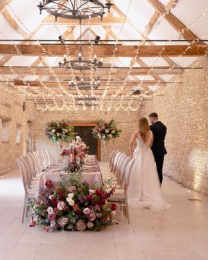 Bride and groom admire a floral-adorned reception table in a fairy-light-filled stone barn venue.
