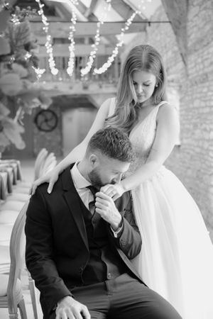 Black and white photo of groom kissing bride's hand while seated in a fairy-lit wedding venue.