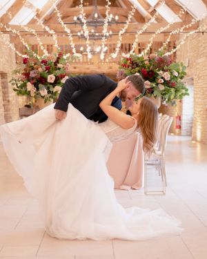 Groom dips bride for a romantic kiss in a rustic barn venue adorned with fairy lights and flowers.