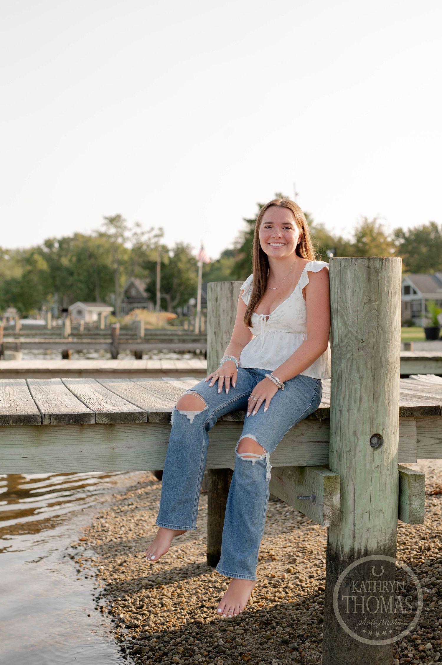 Person sitting on a wooden pier post by the water wearing distressed jeans and a white top.