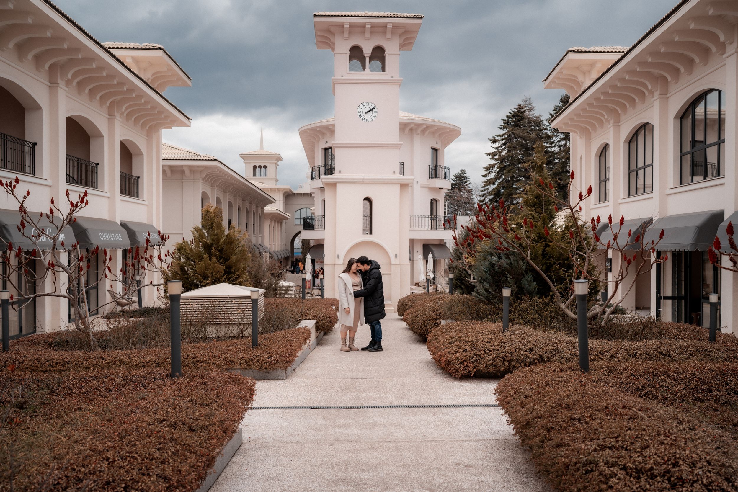 Architectural maternity photo with couple standing on path leading to pink bell tower and white colonnade.