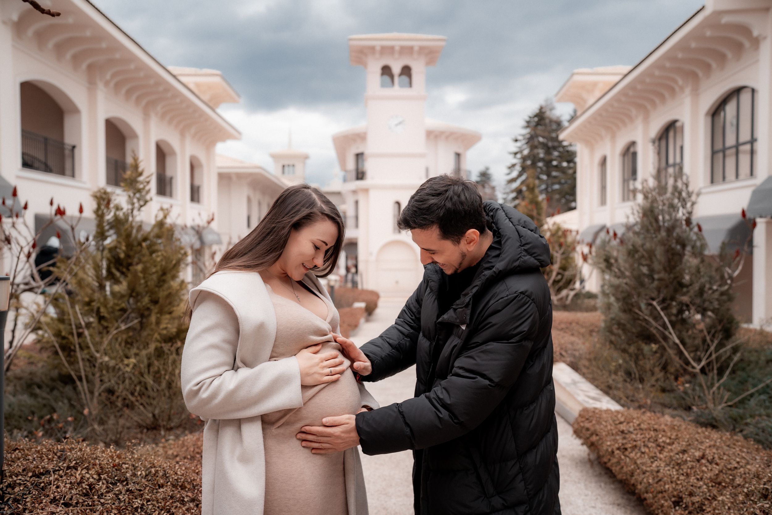 Couple shares tender moment near a Mediterranean-style building with white walls and pink tower during maternity photoshoot.
