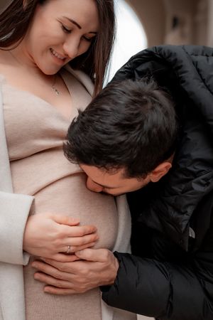 Tender moment as partner kisses pregnant belly while couple poses in soft winter light.