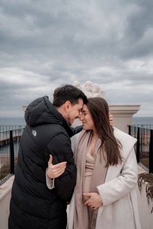 Romantic couple embracing under stormy sky on Mediterranean-style balcony overlooking water.