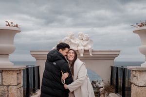Couple sharing intimate moment on coastal terrace with decorative stone pillars and cloudy backdrop.