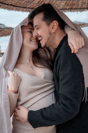 A couple wrapped in a blanket share an intimate moment while smiling and embracing.
