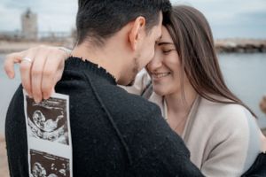 Happy couple sharing a tender moment while holding an ultrasound photo outdoors near water.