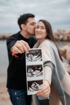 Couple holding ultrasound photos while sharing a sweet moment by the water.