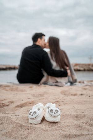 Tiny panda baby shoes on sand with a couple sitting by the water in the background.