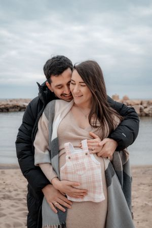 Couple embracing on beach while holding baby clothes, sharing an intimate pregnancy moment.