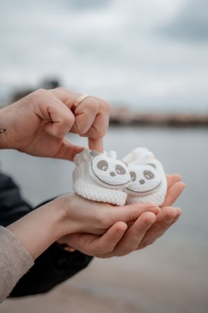 Hands holding white panda baby booties against blurred waterfront background.