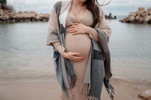 Pregnant woman in beige dress and gray shawl stands by coastal rocks cradling her baby bump.