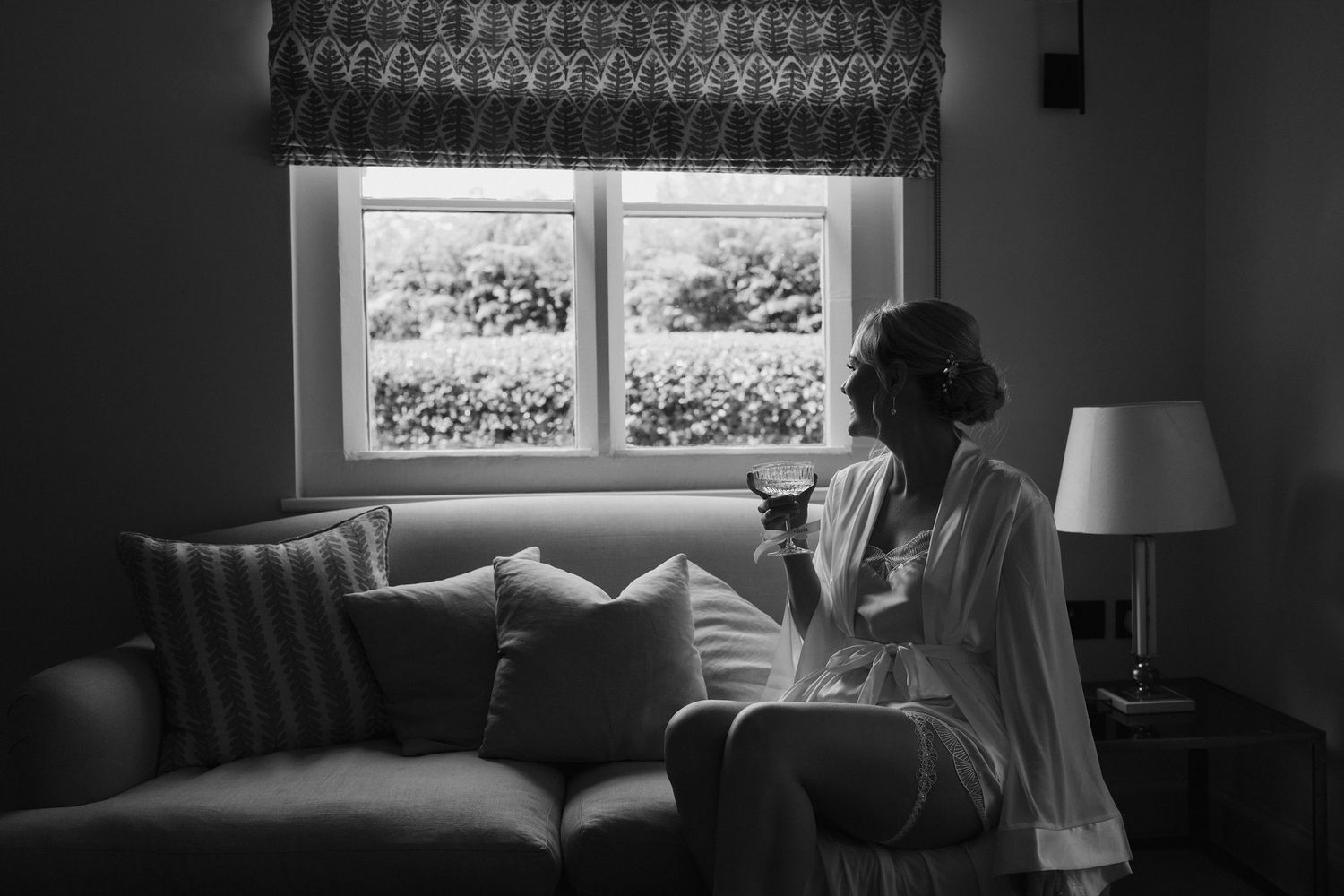 bride sat on sofa looking out window in atmospheric light, holding a champagne glass in her bridal robe at Iscoyd park