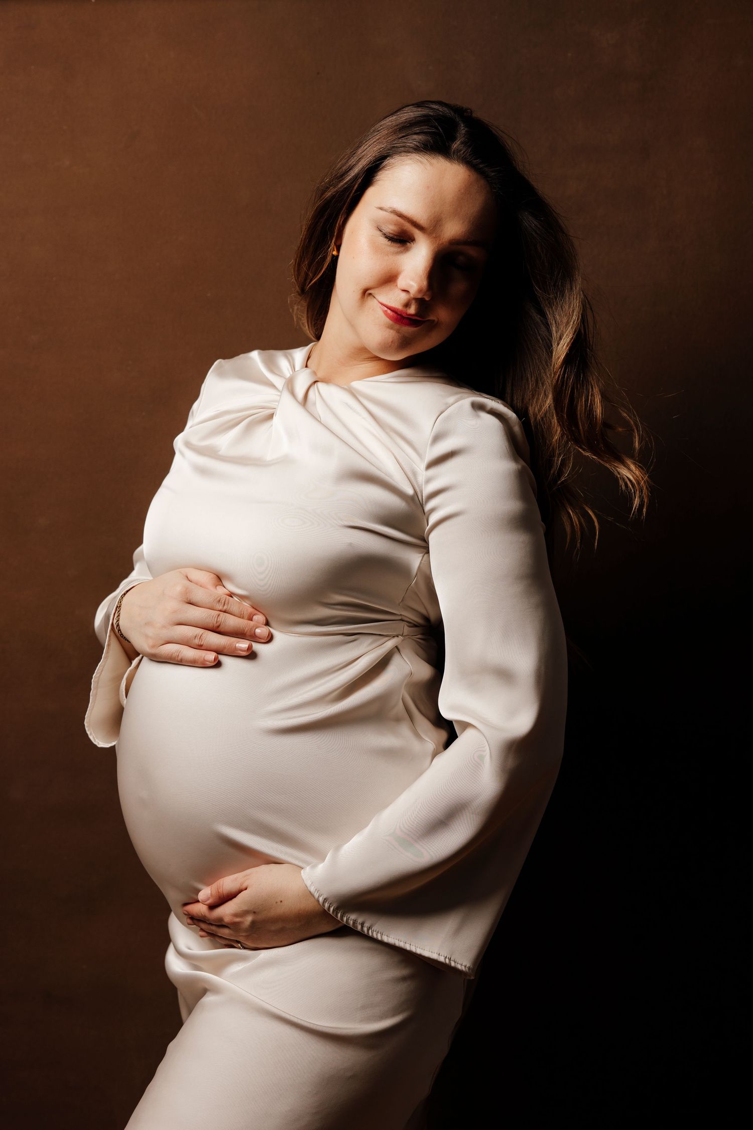 Maternity portrait of woman in white dress against dark background, holding her baby bump.