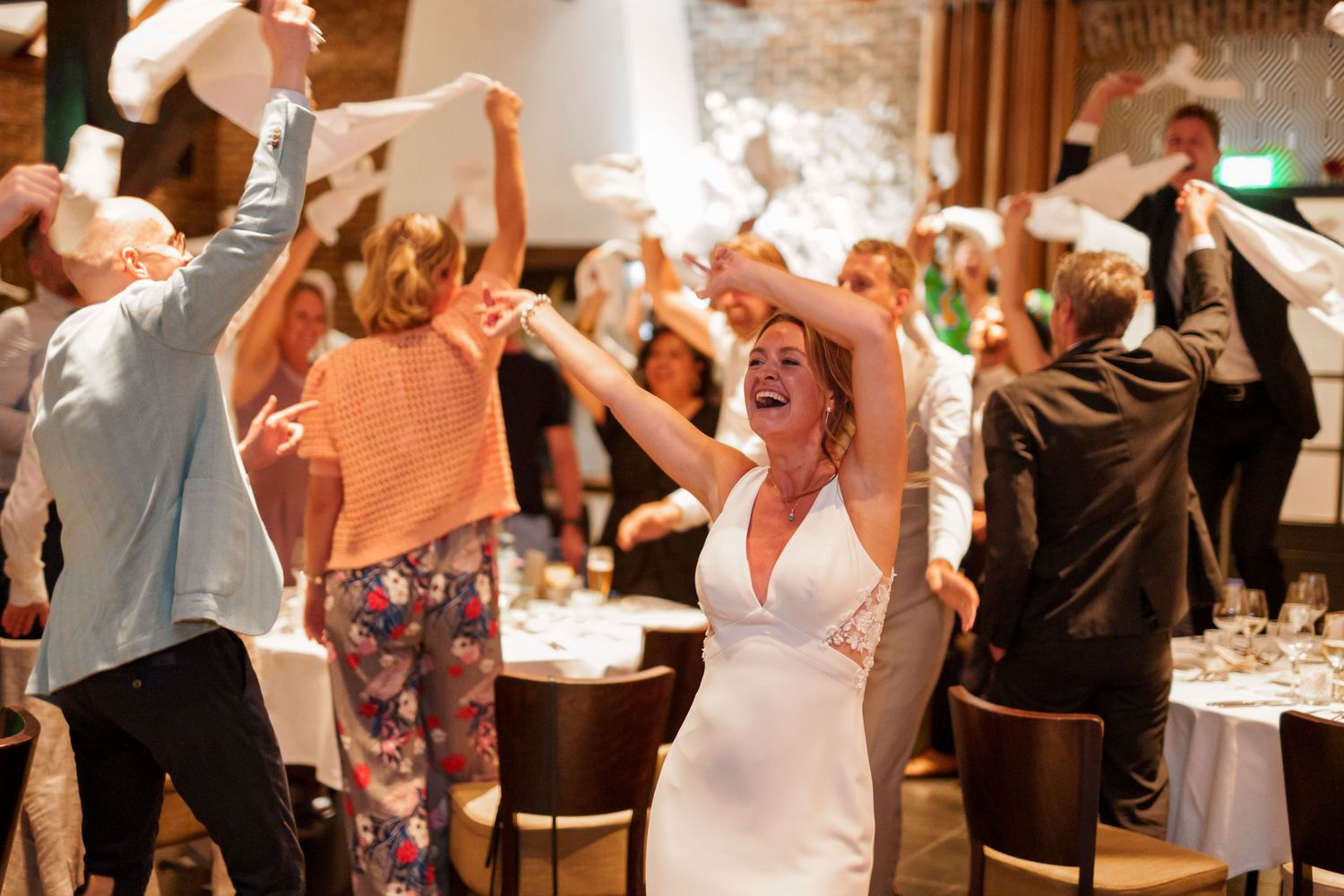 Wedding reception guests dance and celebrate with joyful energy at an indoor venue with warm lighting.