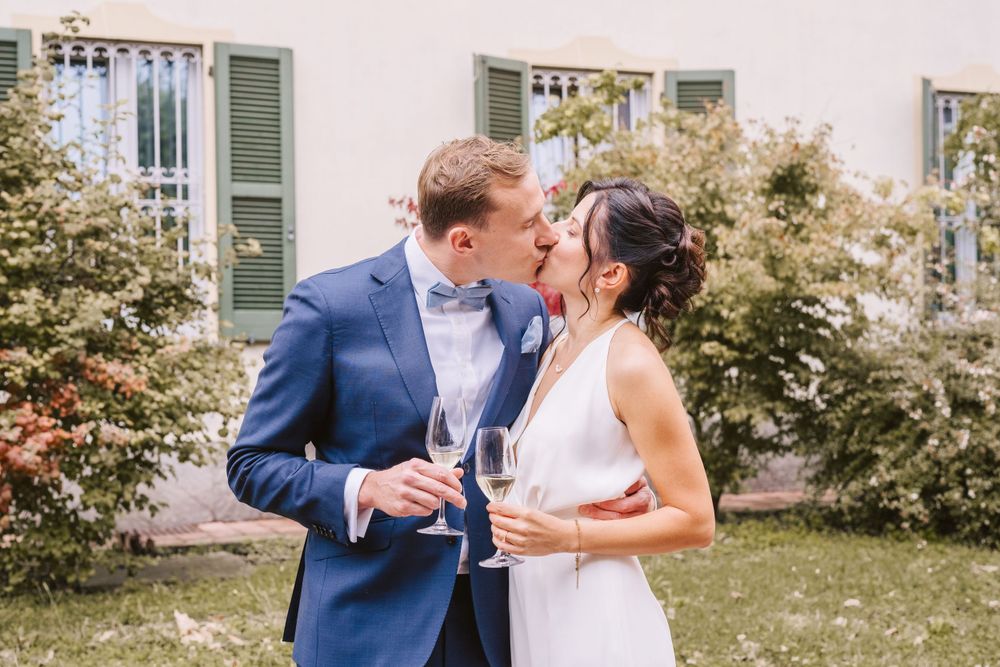 Coppia di sposi si cambia un bacio dopo la cerimonia tenendo un mano due calici di vino