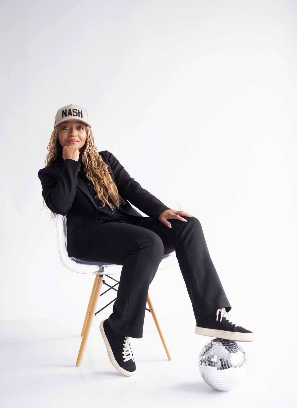 Person in all black attire sits on modern wooden chair against white backdrop in artistic poses.