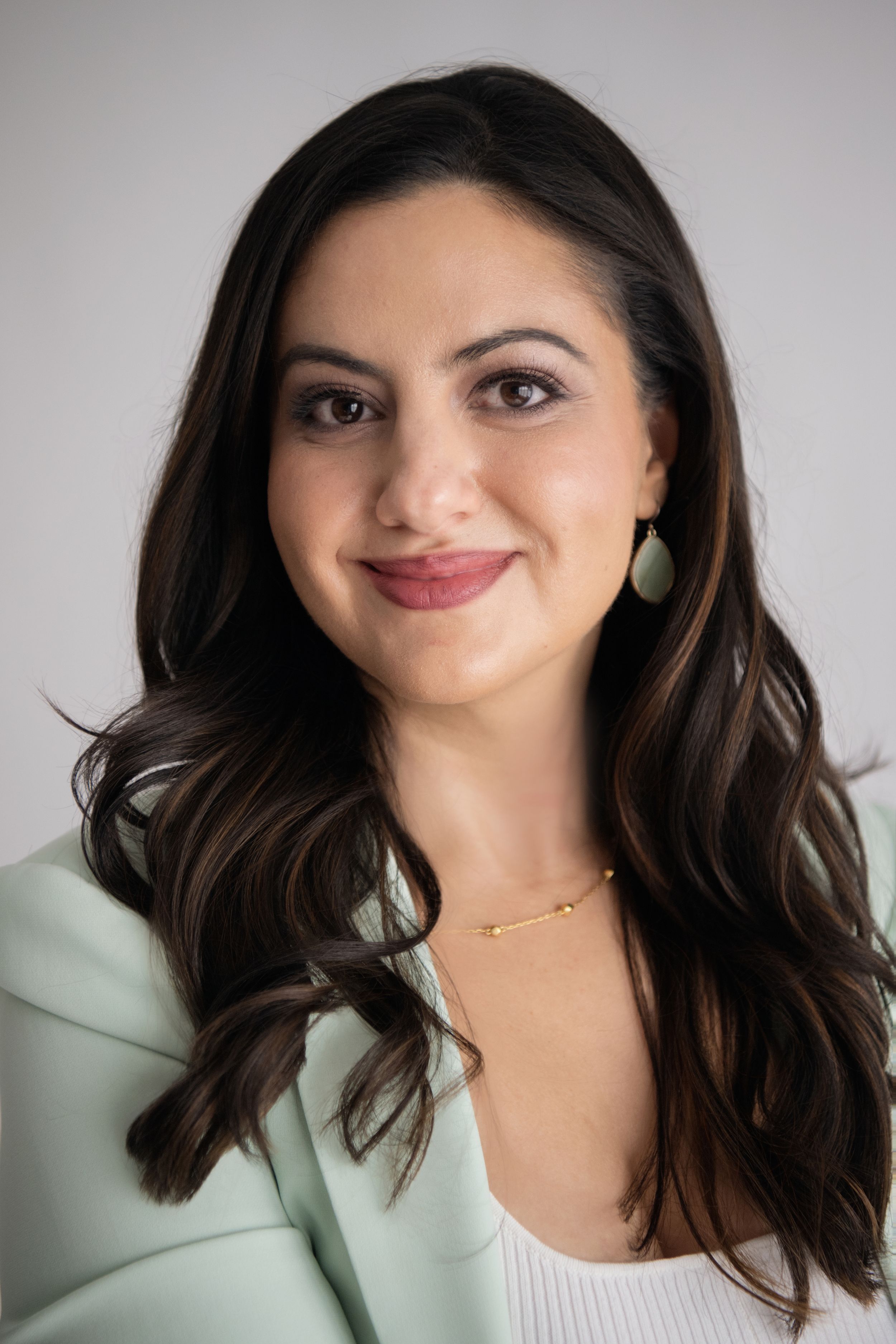 Person with long wavy dark hair and a mint green top smiles at camera in a headshot photo series.